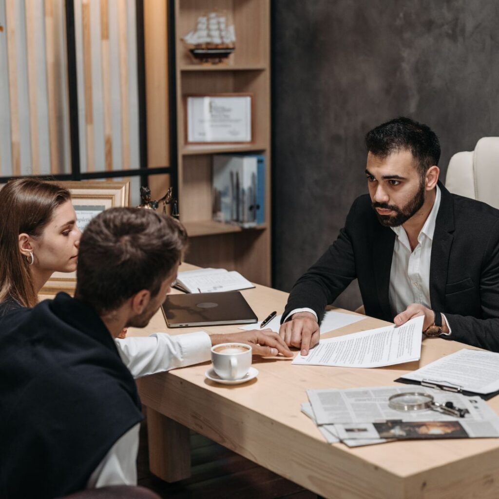 A man in a suit jacket explains a contract to a couple at a wooden desk in an office.